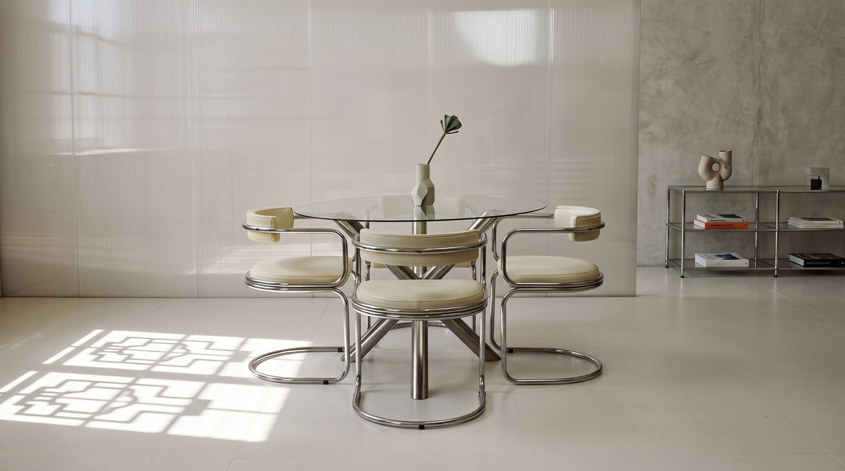 A mid-century modern dining set featuring a round glass-top table and four matching chairs with cream upholstery and chrome frames, set in a minimalist, brightly lit room.